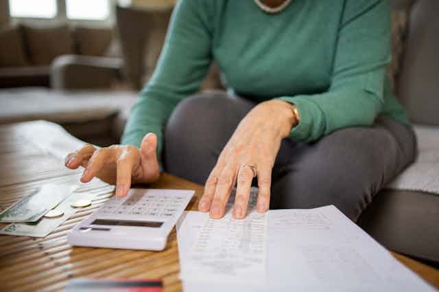 A woman is seen paying bills at a table, with a calculator, bills and receipts around her.