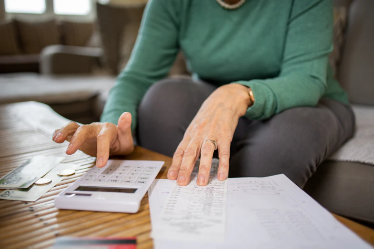 A woman is seen paying bills at a table, with a calculator, bills and receipts around her.