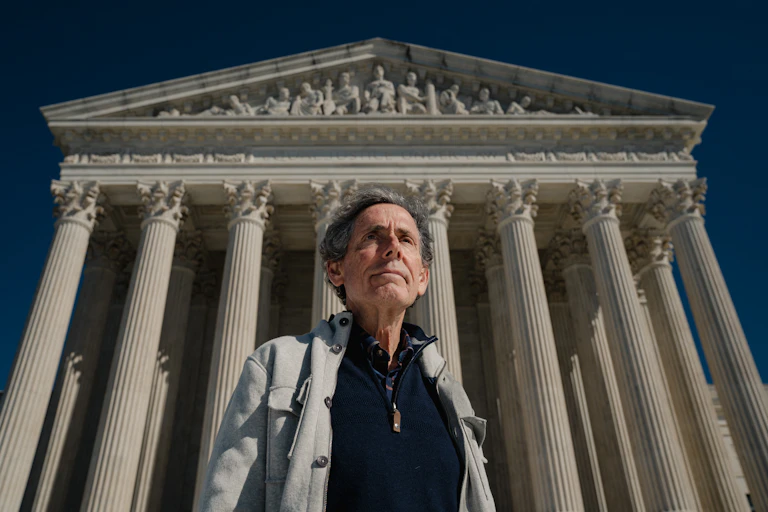 A middle-aged white man stands in front of a white building.