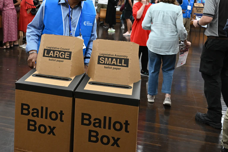 Two ballot boxes are in the centre of a large room. A man in a volunteer vest stands with them.