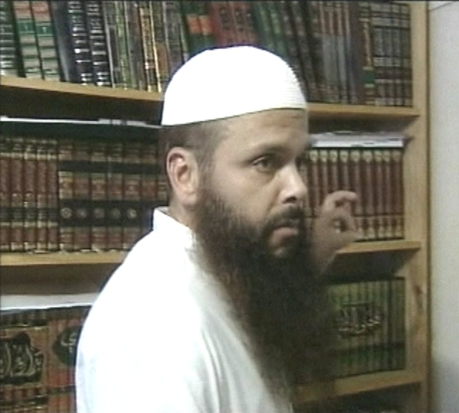 A man in a white gown and hat and long beard stands in front of a bookshelf