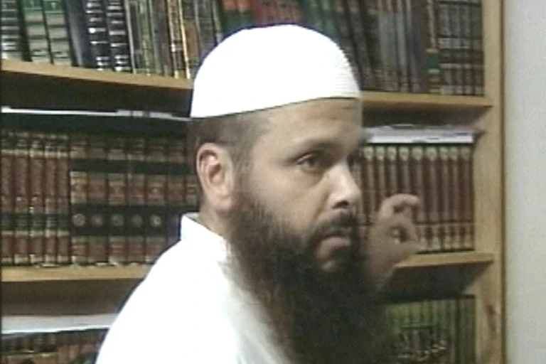 A man in a white gown and hat and long beard stands in front of a bookshelf