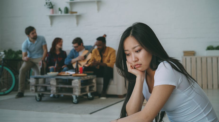 A young women sits away from a group of people, looking sad. 