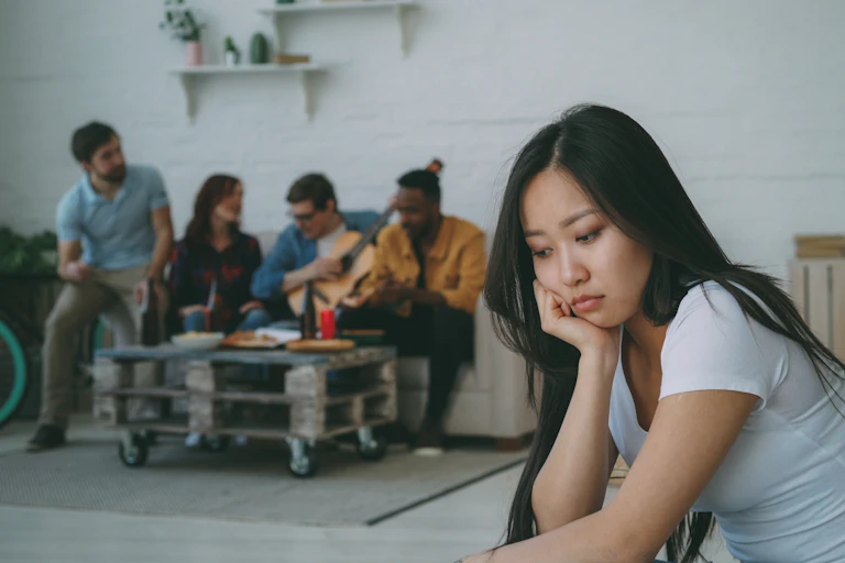 A young women sits away from a group of people, looking sad.