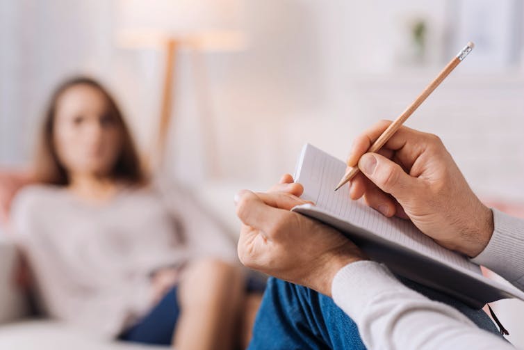 A therapist writes in a notebook while a female patient sits across from them.