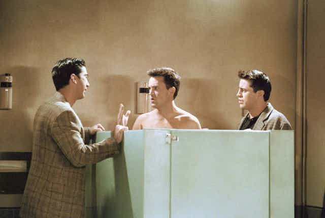 Tres hombres hablan por encima de las puertas de unos baños en un bar.