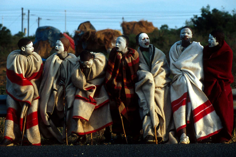 Seven boys in a row, draped in red and white blankets, their faces smeared with white clay and leaning on walking sticks.