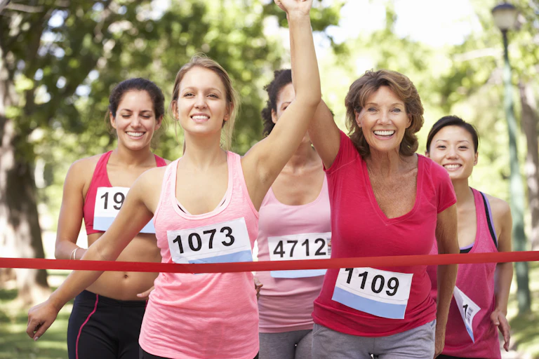 Des coureuses de différents âges, habillées en tenues de sport de couleurs roses et fuschia, et portant des dossards avec différents numéros, sont en train de passer la ligne d'arrivée d'une course. Les deux premières ont joint leur main et l'ont levée.
