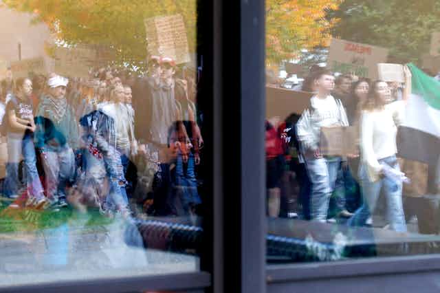A young woman sitting inside looking through a window at a group of people marching.