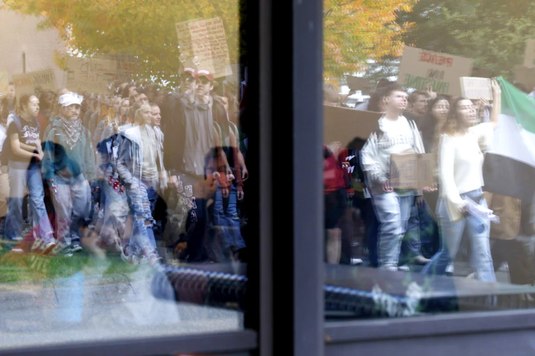 A young woman sitting inside looking through a window at a group of people marching.