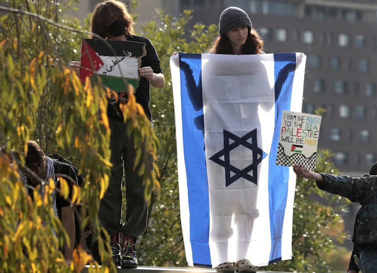 Two people, one holding a Palestinian flag and the other an Israeli flag, with a third person holding a sign in front of the Israeli flag.