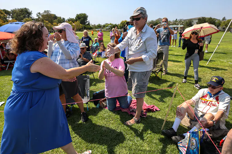 Two people reach their hands out to each other to shake hands in a grassy area with other people around.