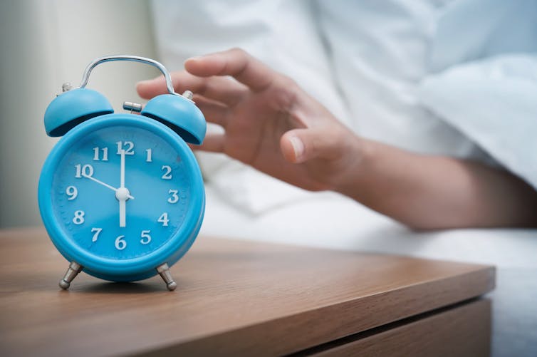 Hand reaching out under bedsheets towards a blue alarm clock on a nightstand