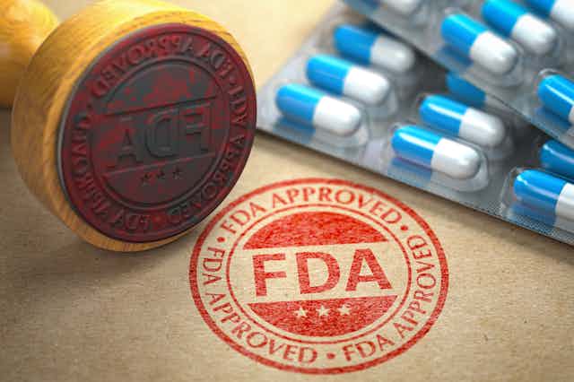 A rubber stamp that reads "FDA approved" is seen in front of a blister pack of pills.