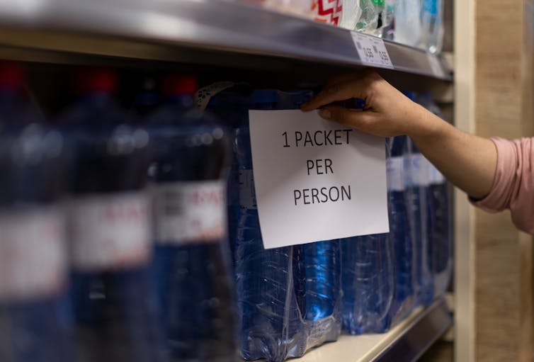 A sign that says '1 packet per person' taped to a shelf of water bottles