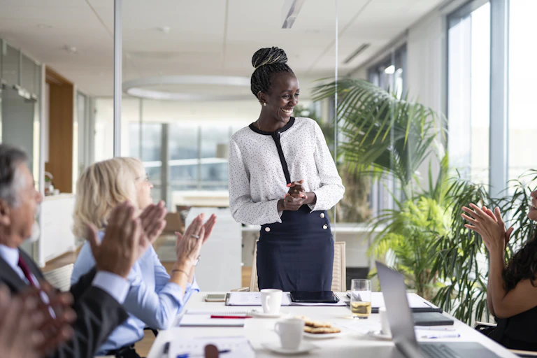 A Black woman smiles while standing at the head of a conference table full of people