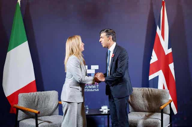 Rishi Sunak and Giorgia Meloni clasp hands and look at each other in front of their respective national flags and two armchairs.