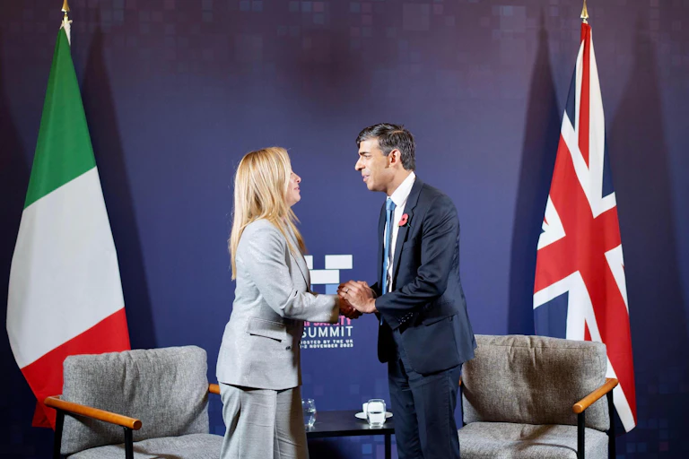 Rishi Sunak and Giorgia Meloni clasp hands and look at each other in front of their respective national flags and two armchairs.