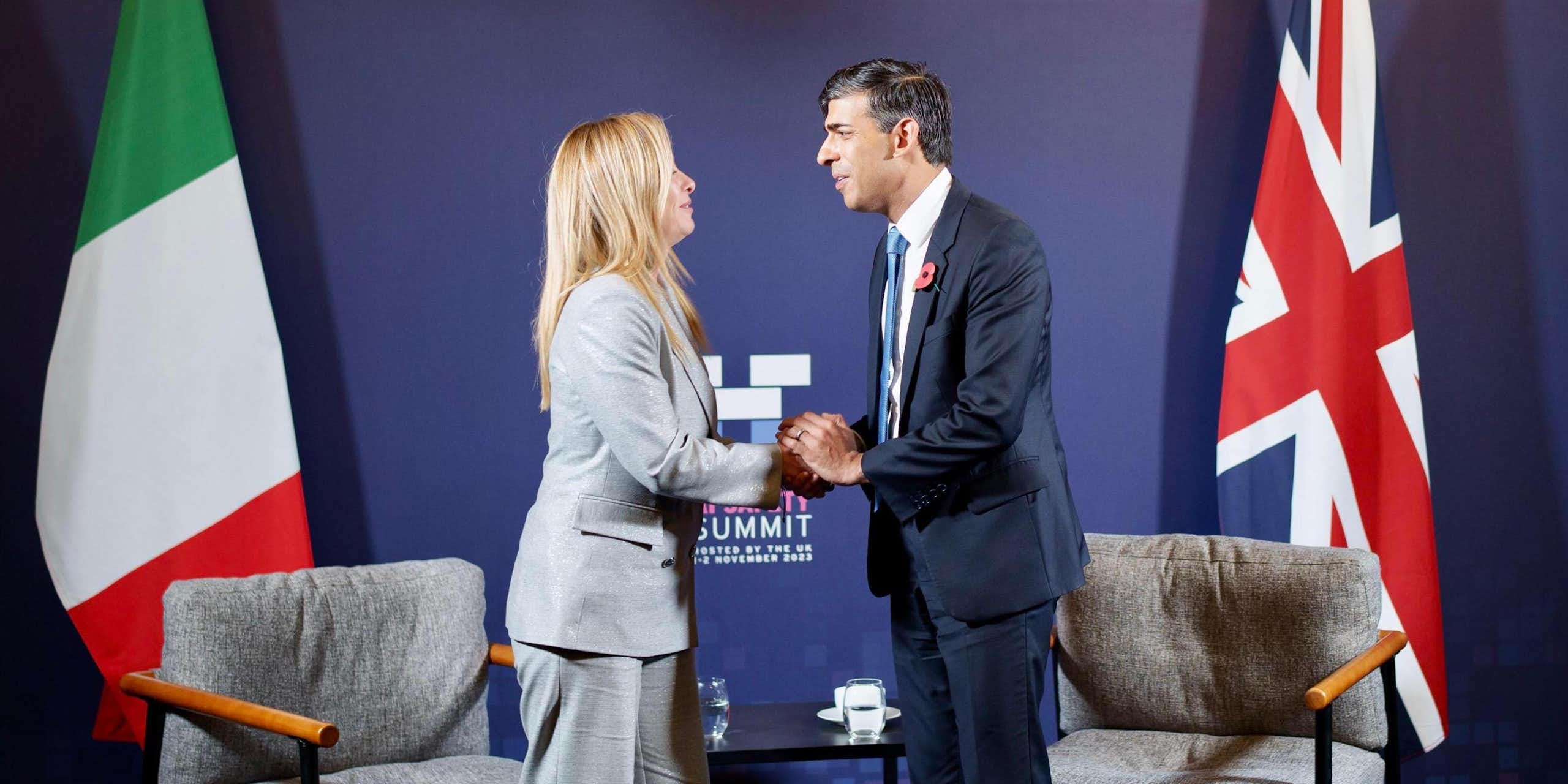 Rishi Sunak and Giorgia Meloni clasp hands and look at each other in front of their respective national flags and two armchairs.
