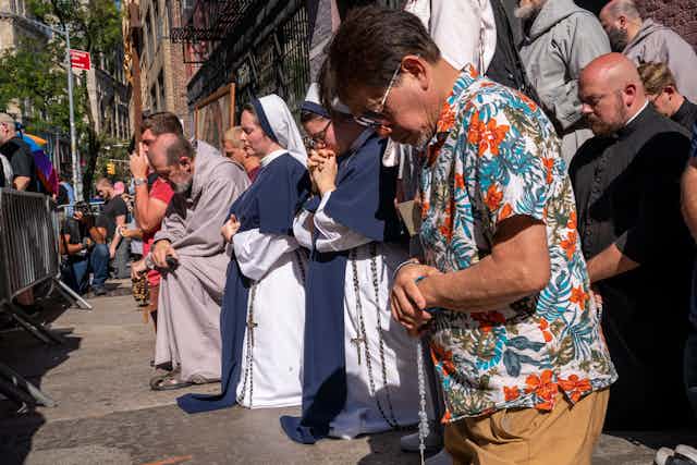 Nuns wearing blue and white crouch on the ground and pray alongside men wearing black and grey robes and a man in a Hawaiian print shirt