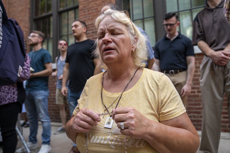 A woman wearing a yellow shirt holds a rosary around her neck and appears to pray with her eyes closed.