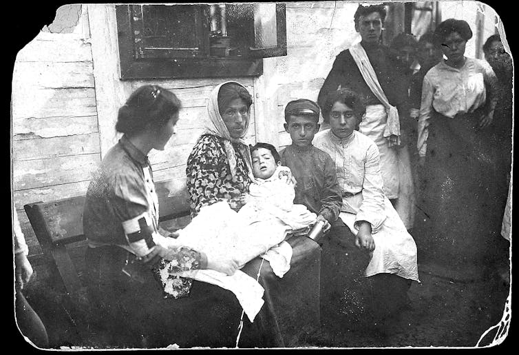 A young injured girl surrounded by her family and attended to by a red cross nurse.