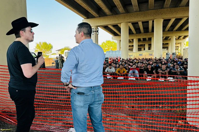 Elon Musk wearing a cowboy hat stands with another man in front of a large group of migrants seated on the ground.
