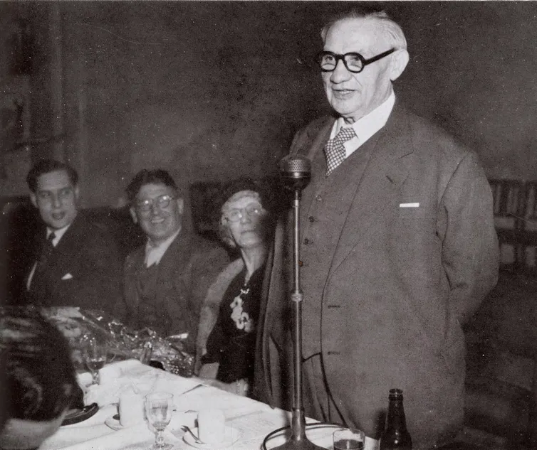 Ernest Bevin speaking into a microphone at a table set for dining with supporters looking on behind him.