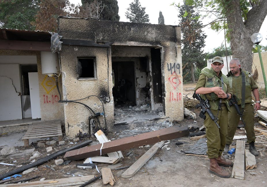 Two Israeli soldiers wearing green and carrying weapons stand outside a bomb damaged house.
