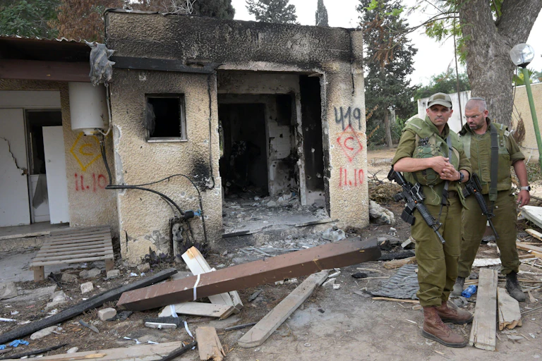 Two Israeli soldiers wearing green and carrying weapons stand outside a bomb damaged house.