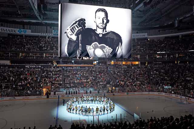 A black-and-white photo of a hockey player is projected on a screen above a hockey rink. On the ice, hockey players stand in a ring.