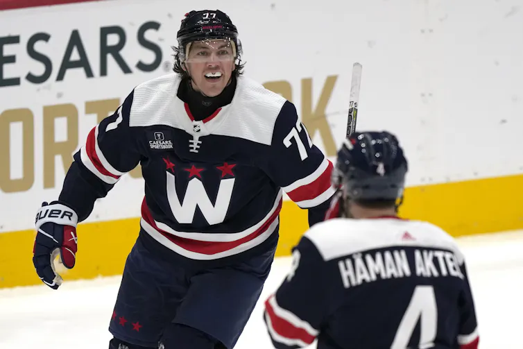 A hockey player wearing a neck guard skates toward his teammate, who has his back to the camera