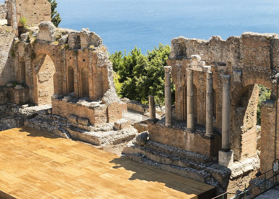 A shiny wooden stage incorporated with ancient, columned ruins, with an ocean in the background.