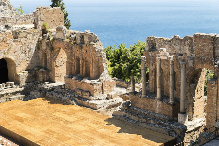 A shiny wooden stage incorporated with ancient, columned ruins, with an ocean in the background.