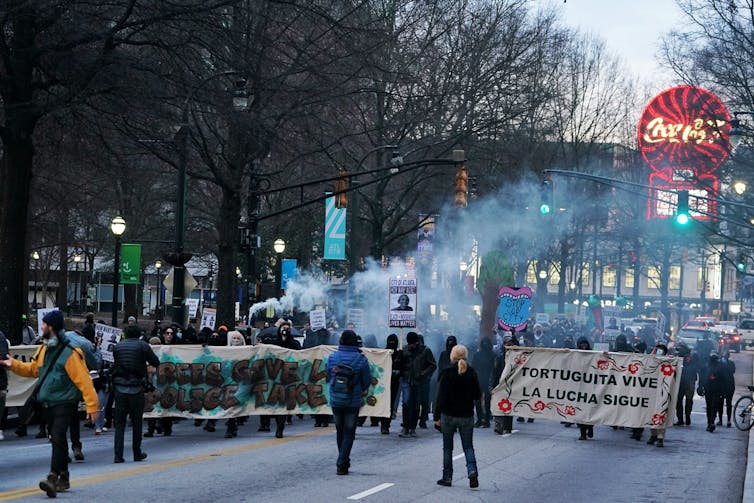 Los manifestantes que portan grandes carteles marchan entre el humo y debajo de un letrero de neón de Coca-Cola.