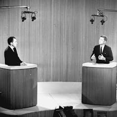 Two men stand at podiums in a black-and-white photo.