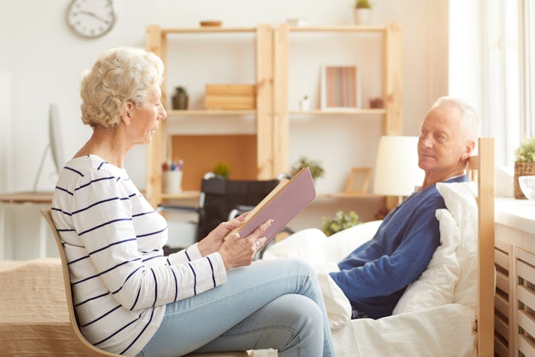 A woman reads to a man sitting up in bed.