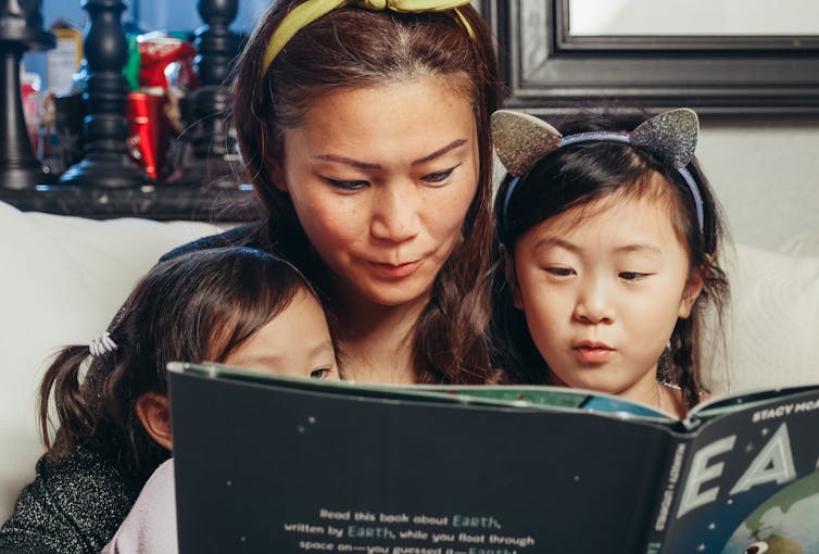 A mother and two young children sit together reading a book.