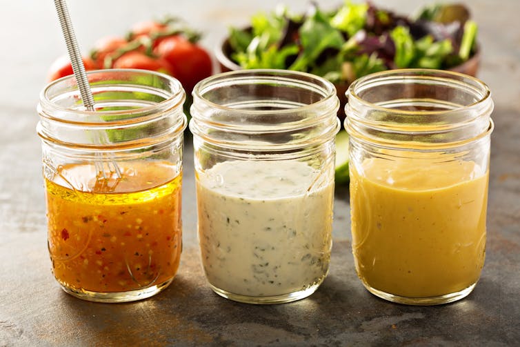 Three salad dressings sit on a bench; one with chilli seeds, one creamy yoghurt-based dressing and one mustard and oil emulsion.