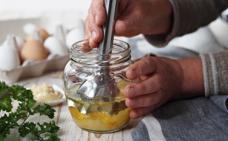 A person makes mayonnaise using a mechanical stick blender.