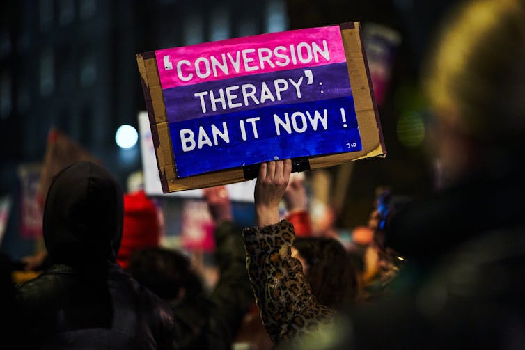 A protester's hand holds a handmade sign reading 'conversion therapy' ban it now!