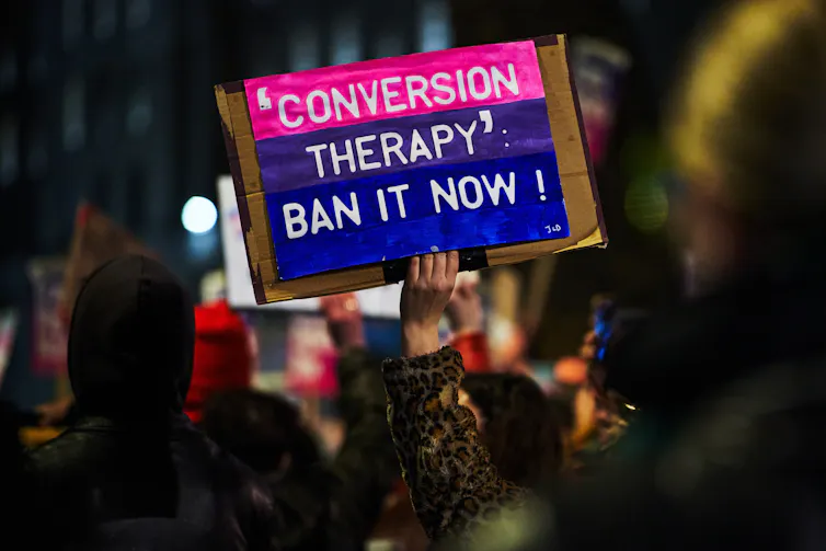 A protester's hand holds a handmade sign reading 'conversion therapy' ban it now!