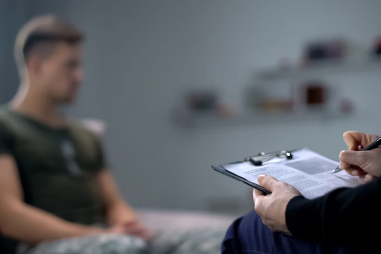 A therapist taking notes on a clipboard, while a young man, blurred in the background, sits on a couch