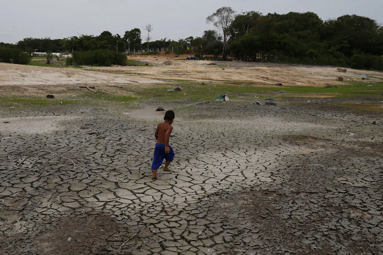 Menino caminha sobre chão seco e quebradiço, que um dia foi o leito de um grande rio