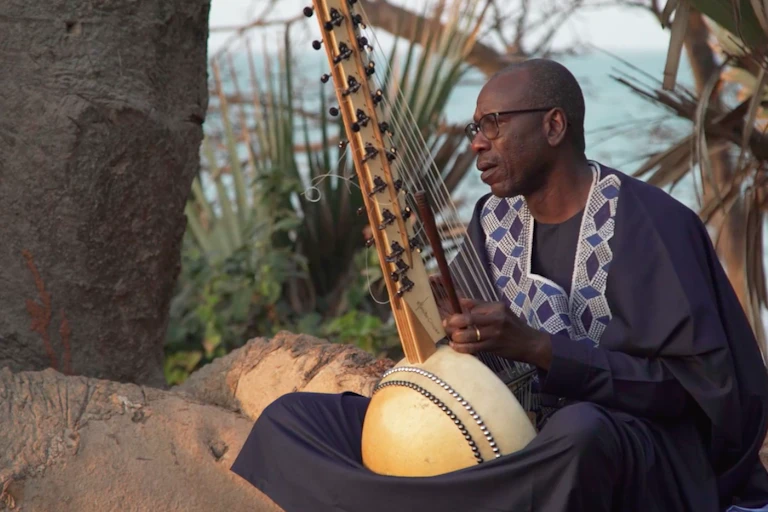 A man sits under palm trees in a flowing traditional west African robe playing a wooden stringed instrument with a bulbous bottom and long stem.