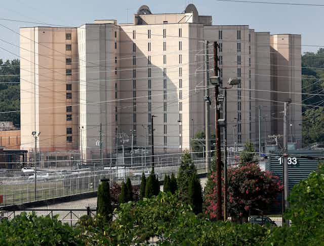 A tall building  has narrow windows and is surrounded by thick metal fencing.