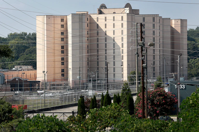 A tall building has narrow windows and is surrounded by thick metal fencing.