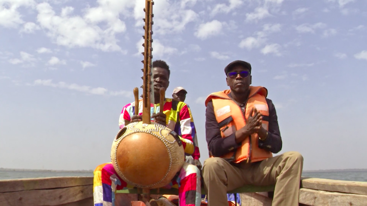 Two men in a boat, one in colourful African print playing a wooden string instrument, white clouds in the blue sky above them.