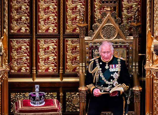 King Charles III, then Prince of Wales, sitting in uniform in an ornate, gilded chair, with the imperial state crown on a cushion beside him