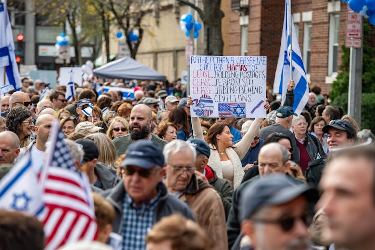 A large group of people walk in the street together, holding Israeli and American flags and a sign that says, 'Rather than a ceasefire, call for Hamas to cease holding hostages, cease terrorism, cease hiding behind civilians.'
